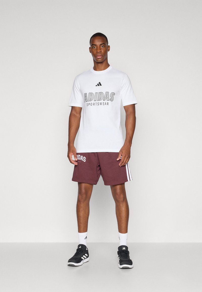 Hombre de pie que lleva puesta una camiseta blanca de Adidas, pantalones cortos burdeos de Adidas con rayas, calcetines blancos de Adidas y zapatillas negras de Adidas sobre un suelo gris.