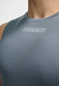 Grå ærmeløs atletisk top med et tekstureret stof, der har et hvidt "HUMMEL" logo på fronten. Nærbillede af halsudskæringen og design detaljer.