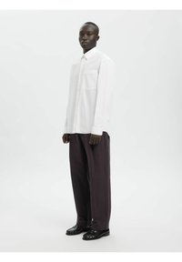 Weißes Hemd mit Knöpfen und vertikalen Streifen, einzelne Brusttasche, kombiniert mit locker sitzenden dunkelgrauen Hosen und schwarzen Schuhen.