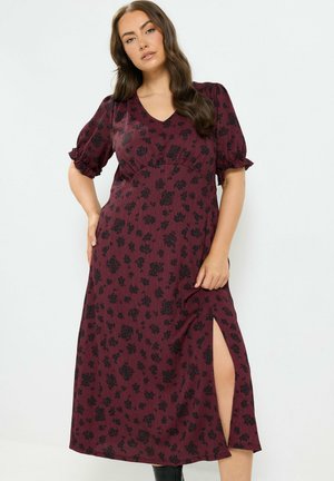 Mujer con un vestido midi burdeos con estampado floral negro, mangas cortas abullonadas y una abertura lateral hasta el muslo, posando con una mano en la cadera.