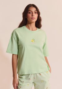 Mujer joven con cabello largo y oscuro que lleva una camiseta verde claro con un gráfico de limón y pantalones cortos a juego con diseños de frutas.