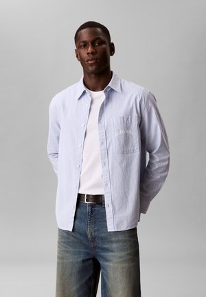 Mann trägt hellblau gestreiftes Hemd über weißem T-Shirt mit blauer Jeans und schwarzem Gürtel und steht vor schlichtem grauem Hintergrund.