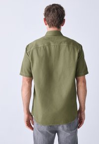 Short-sleeve olive green shirt with textured fabric, a classic collar, and a relaxed fit. Features side slits and a curved hem design.