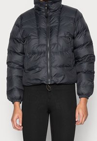 Piumino nero con colletto alto, chiusura con zip, design trapuntato, polsini elasticizzati e orlo regolabile con cordino. Presenta una piccola tasca con zip.