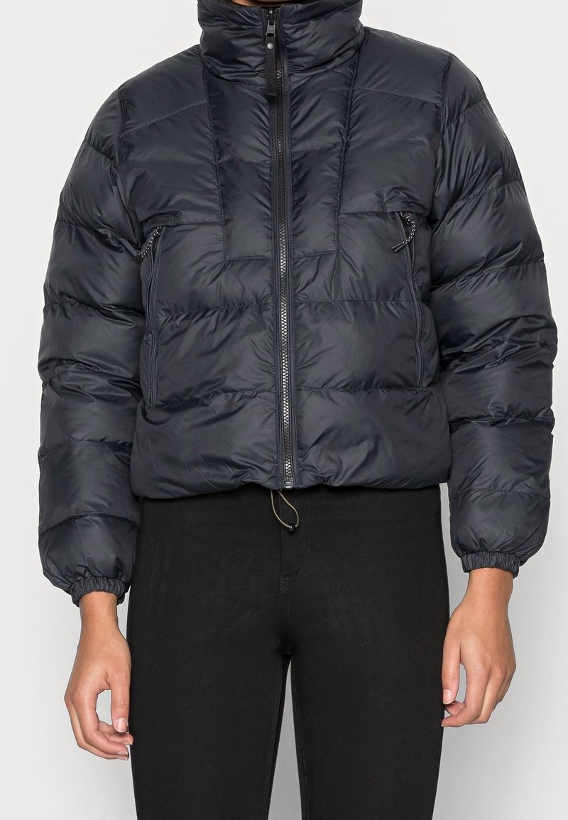 Piumino nero con colletto alto, chiusura con zip, design trapuntato, polsini elasticizzati e orlo regolabile con cordino. Presenta una piccola tasca con zip.