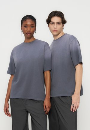 To stående modeller som har på seg matchende grå oversized t-skjorter og mørke bukser, vendt fremover mot en ensfarget hvit bakgrunn.