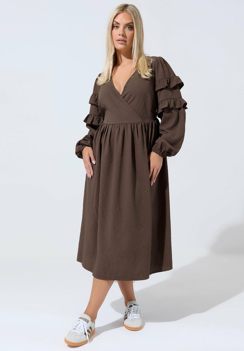 Blonde Frau in einem braunen Midikleid mit langen, gerafften Ärmeln, steht mit übereinandergeschlagenem Bein und trägt weiße Turnschuhe.