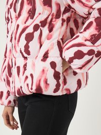 Mulher a usar uma casaco puffer com padrão abstrato em rosa e bordeaux, com uma mão no bolso, combinado com calças pretas.