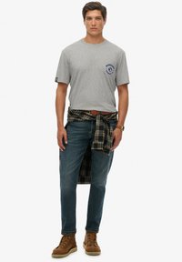 Camiseta gris de manga corta con logo azul, camisa de cuadros azul atada a la cintura, jeans azules y botas marrones con cordones.