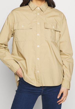 Person wearing a beige button-up shirt with two flap chest pockets and dark blue jeans, hand in pocket, standing against a plain background.