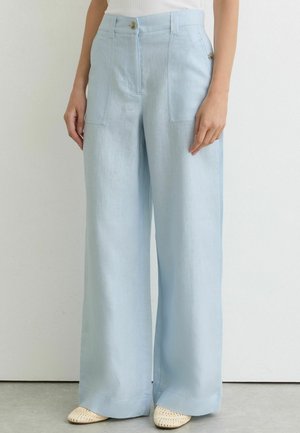 Lichtblauwe wijde broek met voorzakken en knoopdetail, gedragen met witte geweven schoenen en een wit topje.
