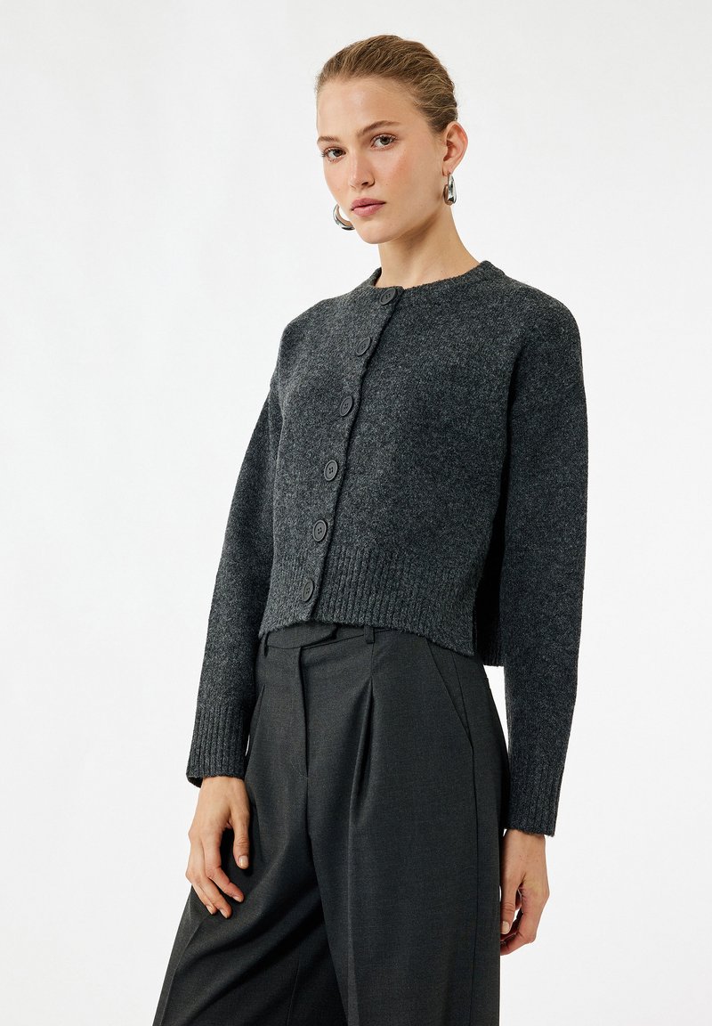 Kvinde iført en mørkegrå knappet cardigan med lange ærmer, parret med højtaljede mørke bukser mod en ensfarvet hvid baggrund.