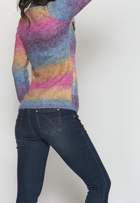 Maglione multicolore a gradiente con tonalità di blu, rosa e arancione; texture in maglia morbida. Abbinato a jeans blu scuro con tasche posteriori decorative.