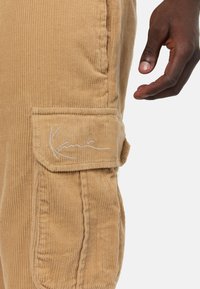 Vaaleanruskeat corduroy-housut sivutaskulla ja brodeeratulla brändilogolla. Housuissa pystysuuntainen ribbineulos ja selkeät saumat.