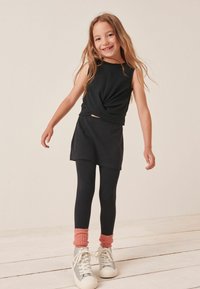 Schwarzes ärmelloses Oberteil mit verdrehtem Front, kombiniert mit einem schwarzen Rock und schwarzen Leggings. Akzentuiert mit rosa Scrunch-Socken und silbernen Sneakers.