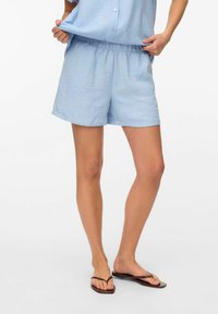 Pantalones cortos de algodón azul claro con cintura elástica. Corte holgado, longitud a medio muslo y combinados con chanclas oscuras. Textura suave.