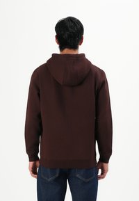 Hombre con cabello negro que lleva una sudadera marrón y jeans azules, de pie de espaldas contra un fondo blanco liso.