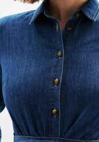 Jeansowa koszula z zapięciem na guziki, kołnierzykiem i brązowymi szwami. Ma brązowe guziki oraz dopasowany fason w talii. Głęboki niebieski kolor.