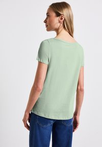 Světle zelený top s krátkým rukávem, volným střihem, kulatým výstřihem a jemnou texturou. V kombinaci s modrými džíny. Minimalistický design.