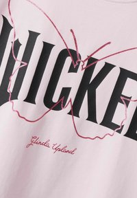 T-shirt en coton rose avec un grand texte noir et un design de papillon rose scintillant. Inclut "Glinda Upland" en cursive en bas.