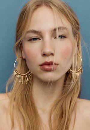 Giovane donna con lunghi capelli biondi che indossa grandi orecchini a cerchio dorati con barre pendenti su uno sfondo blu, che guarda dritto davanti a sé.