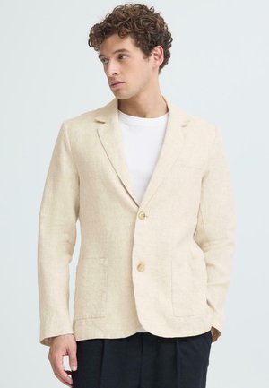 Mann med krøllete hår iført en beige blazer over en hvit skjorte og sorte bukser, ser mot venstre mot en ensfarget bakgrunn.