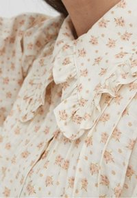 Vit blus med små rosa blommönster, volangkrage och knapprad, tillverkad av lätt tyg med en mjuk yta.