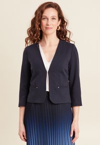 Blazer blu navy sartoriale con scollo a V, maniche tre quarti, tasche laterali con accenti argentati, indossato sopra un top bianco con pizzo e una gonna plissettata.