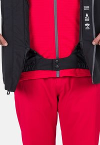 Giacca impermeabile nera con zip grigia, indossata sopra un top rosso texturizzato. Presenta una vita nera regolabile con chiusure a scatto.