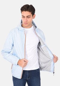 Giacca azzurra chiara con cappuccio, dotata di polsini elasticizzati e chiusura con zip. L'interno è grigio con una tasca, evidenziando un design elegante e minimal.