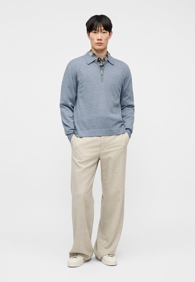Νέος άνδρας που φοράει μπλε πουλόβερ με φερμουάρ πάνω από μια καρό πουκάμισο, μπεζ παντελόνι με φαρδιά πόδια και λευκά αθλητικά παπούτσια, στέκεται με τα χέρια στις τσέπες.