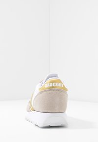 Bela in bež teniska z odtenki iz semiša, podloženim ovratnikom in teksturirano belo podplatom. "Saucony" logotip v belem na rumenem petnem traku.
