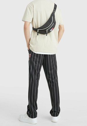 Sac à bandoulière noir avec zippers argentés, design incurvé et sangle réglable, porté sur une chemise beige et associé à un pantalon noir à rayures.