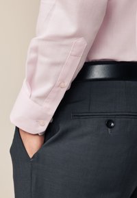 Braccio in manica di camicia da uomo rosa pallido con polsino abbottonato che poggia vicino a pantaloni eleganti grigio scuro con cintura nera e mano in tasca.