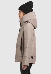 Ljus beige vadderad jacka med hög krage, diagonalt quiltat mönster, dragkedjefickor och en svart stickad mössa. Sido vy visas.