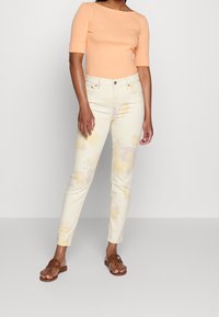 Lehký top z bavlny v broskvové barvě, spárovaný s skinny džíny v květinovém vzoru v žlutobéžové barvě. Oblečení doplňují hnědé ploché sandály.