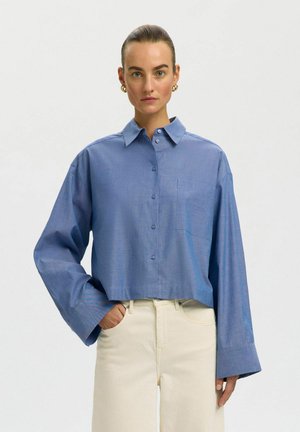 Kvinde med trukket hår tilbage iført en stor blå skjorte med knapper og cremefarvede bukser med høj talje, stående mod en ensartet lyse baggrund.