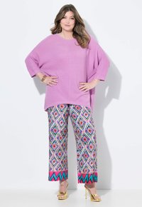 Übergroßer fliederfarbener Pullover mit kurzen Ärmeln, kombiniert mit gemusterten, weit geschnittenen Hosen in lebhaften Farben, die geometrische Muster und pinke Akzente aufweisen.