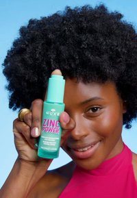 Zinc Power-serum i en teal-flaska med rosa text. Rund pump-topp. Modell med lockigt svart hår, klädd i en rosa topp mot en blå bakgrund.