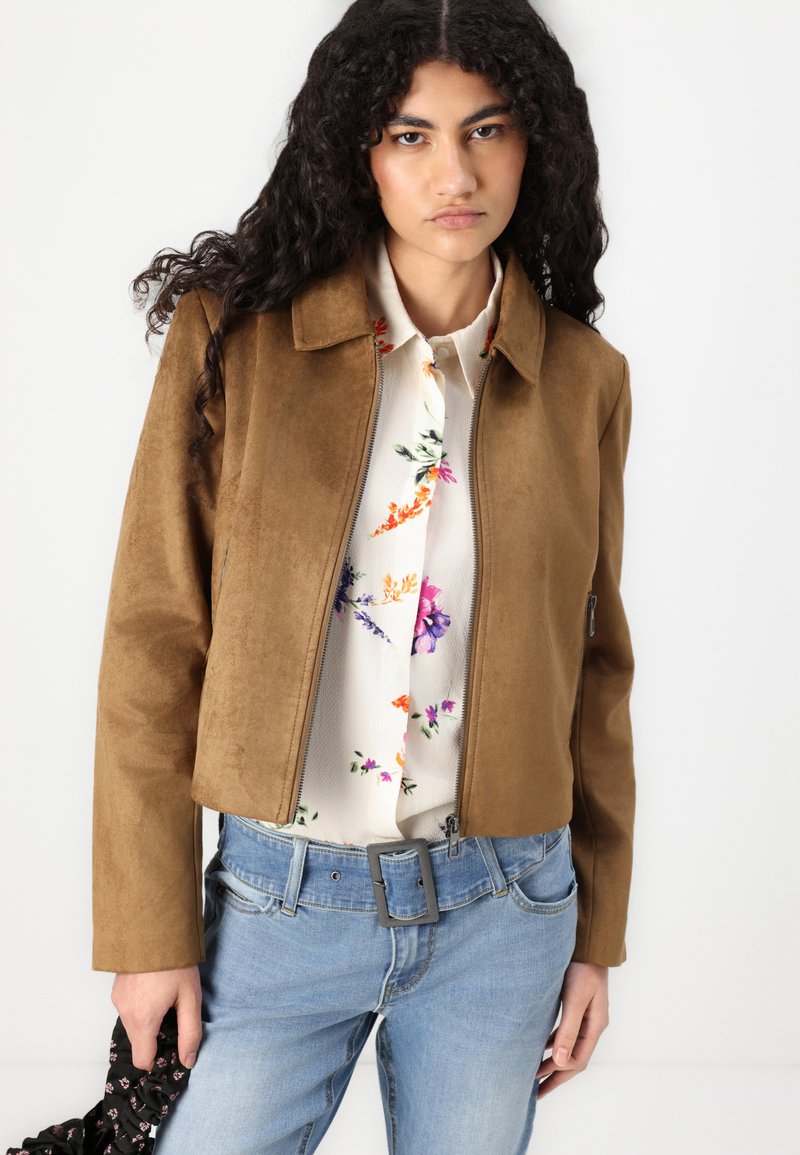 Giacca in suede marrone con chiusura a zip e colletto, indossata sopra una blusa floreale, abbinata a jeans azzurri chiari e un accessorio nero.