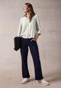 Blusa verde chiaro a fantasia con scollo a V e maniche arrotolate; jeans blu scuro con vestibilità dritta; sneakers bianche; tiene un oggetto nero.