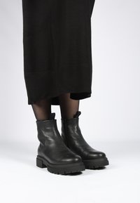 Botas de cuero negras hasta el tobillo con suela gruesa y acabado texturizado, que presentan cremalleras laterales y tiradores para ponerlas y quitarlas fácilmente.