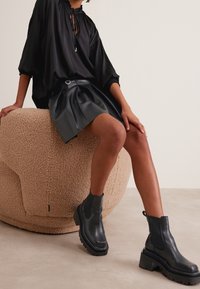 Czarne skórzane buty typu chelsea z masywną podeszwą i ozdobnym szwem, zestawione z luźną czarną bluzką i czarną plisowaną spódnicą z eko skóry.