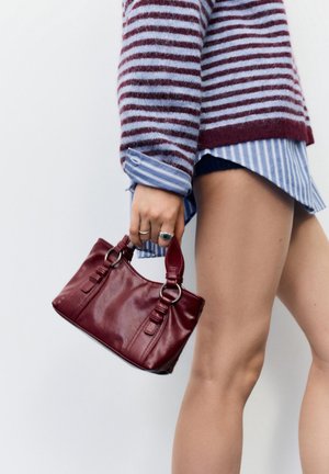 Persoană care poartă pulover cu dungi și cămașă ține o mică geantă de mână din piele culoare burgundy de mânere, arătând picioarele goale pe un fundal neutru.