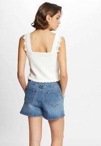 Weißes Tanktop mit Spitzenbesatz an den Trägern, gewelltem Saum, kombiniert mit blauen Jeans-Shorts mit hinteren Taschen und einem Saum, der mittig am Oberschenkel endet.