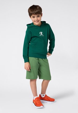 Junge lächelt, trägt einen dunkelgrünen Champion-Hoodie, hellgrüne Shorts, weiße Socken und leuchtend orangefarbene Turnschuhe, stehend vor einem weißen Hintergrund.