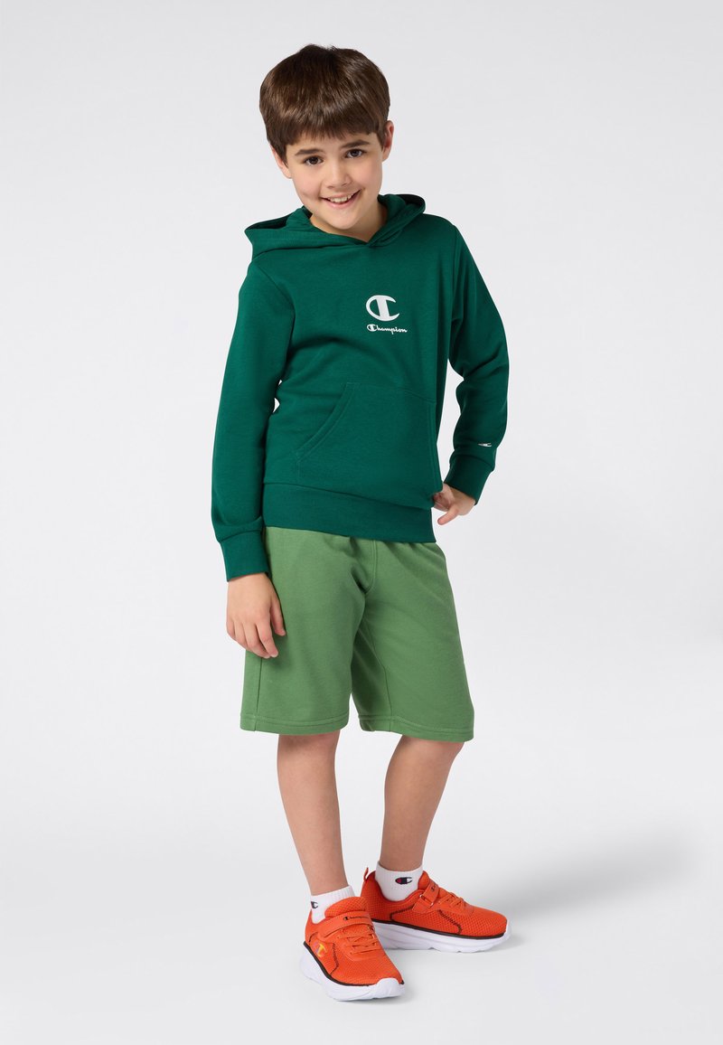 Ragazzo sorridente che indossa un felpa Champion verde scuro, pantaloni corti verde chiaro, calze bianche e sneakers arancioni brillanti, in piedi su uno sfondo bianco.