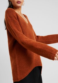 Rostfärgad stickad tröja med V-ringning, långa ärmar och strukturerad tyg. Sidoklyftor och en avslappnad passform framhäver både komfort och stil.