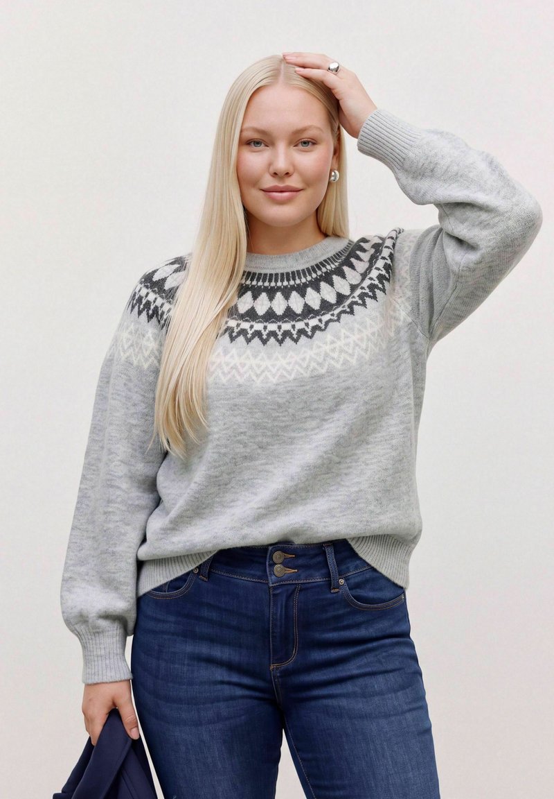 Une femme pose avec un pull gris à motifs et un jean bleu foncé, une main posée sur la tête, devant un fond uni.
