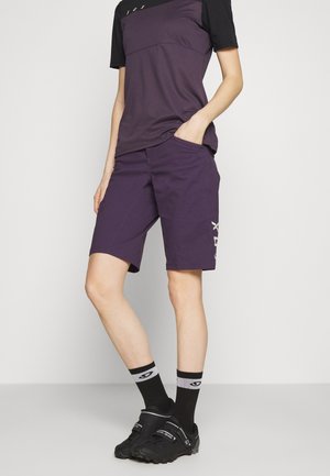 Osoba nosząca fioletowe spodenki rowerowe i pasującą koszulkę, czarne skarpetki z szarymi paskami oraz czarne buty rowerowe, stojąca z jedną ręką w kieszeni.